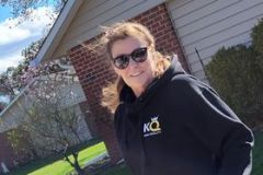 Woman in sunglasses and black hoodie standing outside by a house.