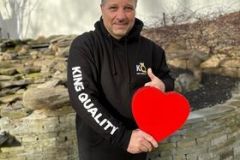 Man holding a red heart outdoors with a slight smile.