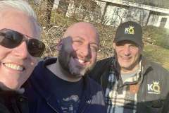 Three men smiling for a selfie in sunny outdoors with a house in the background.