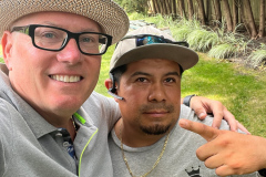 Two men taking a selfie, one gesturing a peace sign, outdoors with greenery.