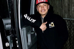 Man in a 'KING' hat and hoodie stands by a vehicle, smiling.