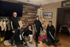 Four people and a dog by a Christmas tree with stockings on the mantel.