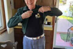 Elderly man smiling and pointing at his KQ T-shirt indoors.