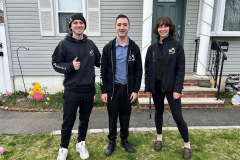 Three young people in KQ jackets smile and give a thumbs-up in front of a house with porch decorations.