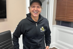 Man in black hoodie and cap smiling in an office setting.