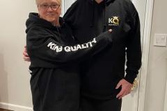 A smiling elderly couple standing close, wearing matching black hoodies.