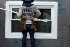 Person installing a window while standing on a stepladder, wearing a cowboy hat and tool belt.