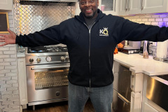 Person in a kitchen with "THE KING" on their jacket, arms spread wide.