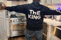 Man smiling with arms outstretched with a "THE KING" sweatshirt on in a modern kitchen.