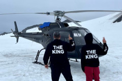 Two people in "THE KING" hoodies next to a helicopter on snow.