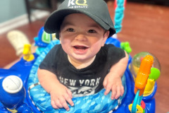 A smiling baby in a blue activity walker wearing a black KQ cap.