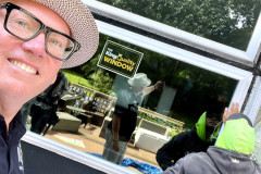 Person taking a selfie with workers installing a window in the background.