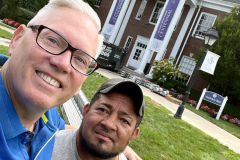 Two men taking a selfie in front of Gaebelein Hall with banners.