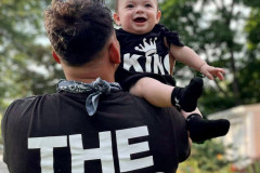 Man with "THE KING" shirt lifting a happy baby wearing "KING" onesie.