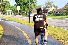 Cyclist with "THE KING" shirt riding on a sunny road near houses.